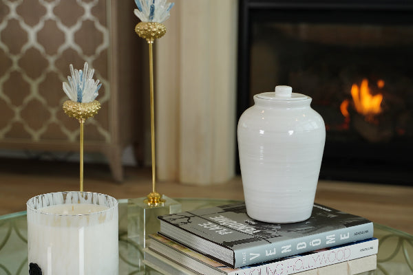 The Modern Classic Cloud Memorial Urn by Stoneware & Co. is elegantly displayed on a glass coffee table, resting atop a curated stack of designer books. This handcrafted ceramic cremation urn, finished in a serene cloud white, blends seamlessly into the sophisticated home décor. The warm glow of a nearby fireplace enhances the urn’s timeless beauty, making it a peaceful and dignified tribute to a loved one. Available through Magnolia Cremations.