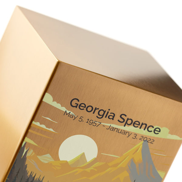 A close-up of the Mountain Landscape in Bronze Memorial Urn by Magnolia Cremations, highlighting the fine brushed metal surface and precise engraving, which is shown as an available option. The urn’s artwork features a golden sunset casting light over majestic mountains, a serene river, and a lush forest, symbolizing tranquility and the beauty of nature. The smooth, polished edges of the urn enhance its refined, contemporary design, making it both a heartfelt tribute and a striking decorative piece.
