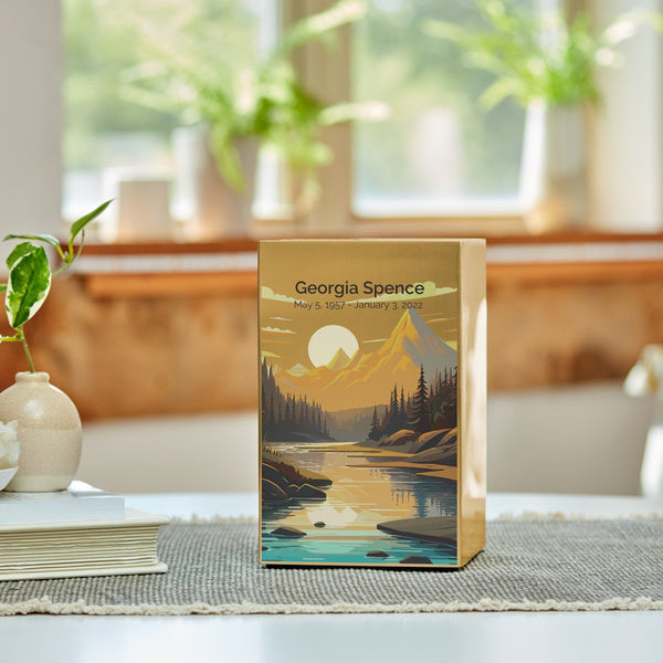 The Mountain Landscape in Bronze Memorial Urn by Magnolia Cremations sits gracefully on a table in a well-lit home, bathed in natural sunlight. This contemporary urn features a stunning nature scene with a golden sunset over towering mountains, a calm reflective river, and a forested landscape, symbolizing peace, adventure, and eternal rest. The brushed bronze finish complements the vivid artwork, making it both a meaningful tribute and a striking decorative piece. Engraving is shown as an available option.