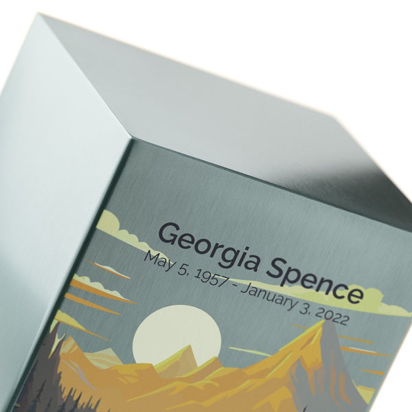 A close-up of the Mountain Landscape in Pewter Memorial Urn by Magnolia Cremations, highlighting its brushed pewter surface and intricate artwork. The design features a peaceful mountain scene with a glowing sun casting warm light over the rugged peaks and a reflective river below. The engraving is shown as an available option, offering a personalized tribute. The smooth, metallic finish catches the light, emphasizing the urn’s elegant craftsmanship.