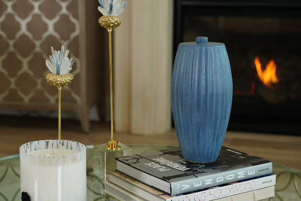 A full-size ceramic urn in sky blue from the exclusive Nouveau Groove collection for Magnolia Cremations by Stoneware & Co. The urn features a gently tapered shape with vertical ridges, inspired by the Art Nouveau style. It is displayed on a glass coffee table, resting on a stack of design books, with a softly flickering fireplace in the background. The urn’s smooth glaze has subtle variations in tone, adding depth and elegance to its contemporary design.