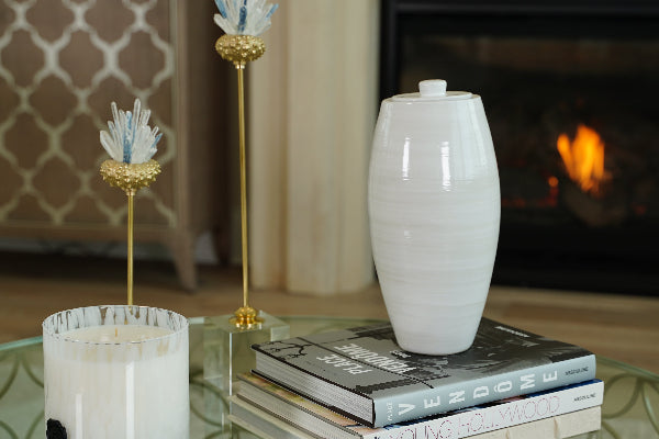 The Nouveau Pure Cloud Memorial Urn, an exclusive design for Magnolia Cremations by Stoneware & Co., is displayed in a serene home setting. The full-size ceramic urn, finished in a soft cloud white glaze, sits atop a stack of elegant books on a glass coffee table. Its smooth, tapered form and minimalist design reflect Art Nouveau inspiration. A fireplace glows warmly in the background, alongside decorative gold and blue floral sculptures, creating a peaceful and dignified atmosphere.