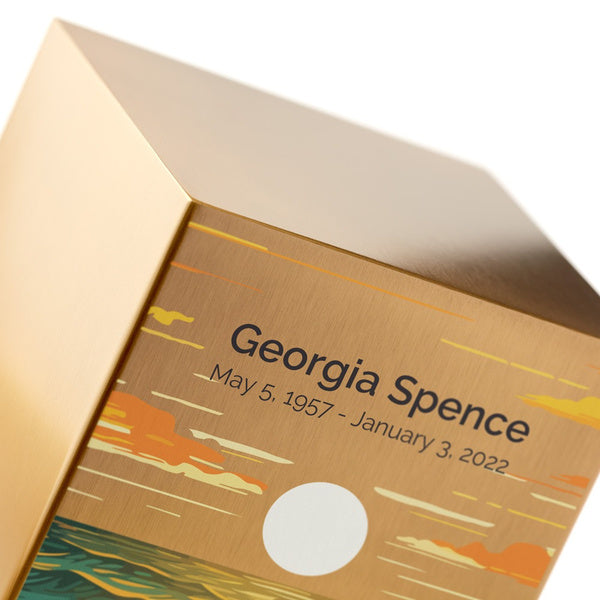 A close-up of the Ocean Landscape in Bronze Memorial Urn, showing its brushed stainless steel surface with a warm bronze finish. The urn features an artistic ocean scene with rolling waves, a glowing sun, and a colorful sky. Engraving is shown as an available option. This urn is available at Magnolia Cremations.