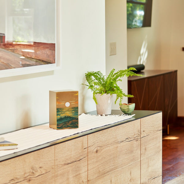 The Ocean Landscape in Bronze Memorial Urn is displayed on a modern wooden sideboard with a black top. The urn features a brushed stainless steel finish adorned with an artistic seascape, depicting rolling ocean waves in deep blue and golden hues beneath a glowing sun. A white cloth runner extends across the surface, with a green fern in a white ceramic pot and a small decorative bowl beside the urn. The background features a warmly lit room with natural light streaming in.
