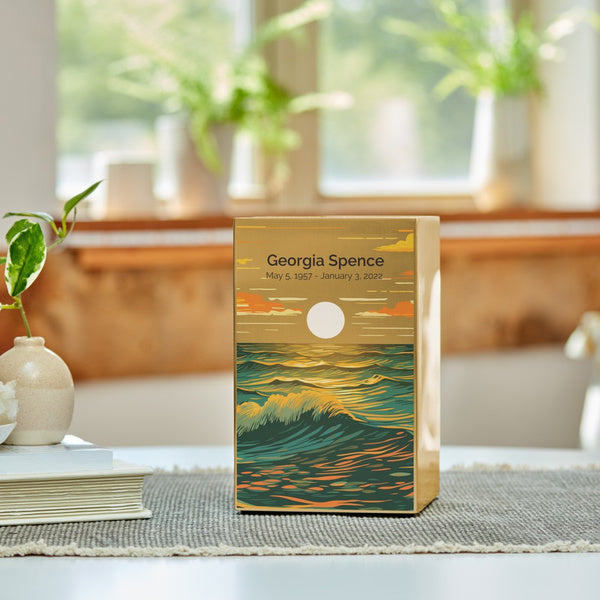 The Ocean Landscape in Bronze Memorial Urn is displayed on a table in a sunlit room, with soft natural light filtering through a window. The urn features a brushed stainless steel finish with an artistic seascape design, showcasing rolling waves in deep blue and golden hues beneath a glowing sun. A woven table runner, books, and a small ceramic vase with a green plant accompany the urn, creating a peaceful and elegant setting. Engraving is shown as an available option.