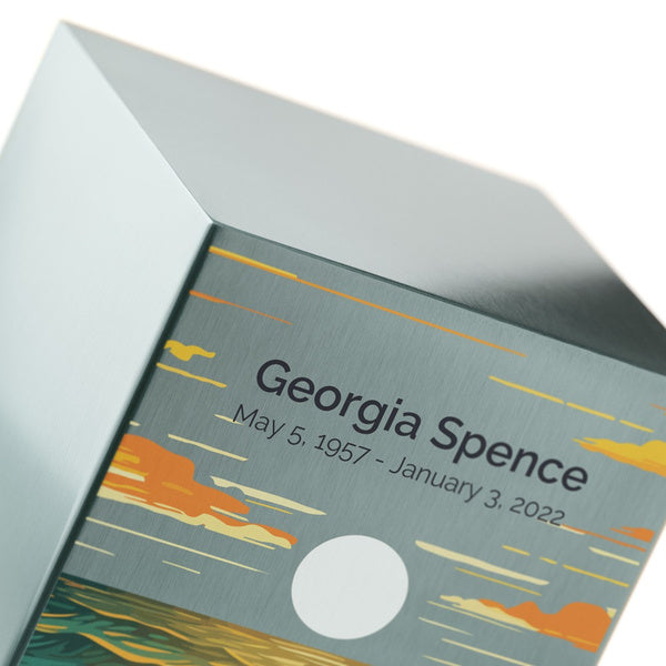 A close-up of a pewter-finished stainless steel memorial urn, highlighting its engraved personalization and vibrant ocean landscape design. The artwork features golden waves illuminated by a full moon, with a sky streaked in warm sunset hues. The brushed metal surface contrasts with the intricate detailing of the design. Personalization is available, making this urn, offered at Magnolia Cremations, a meaningful tribute to a loved one.