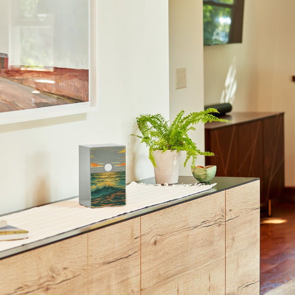 A pewter-finished stainless steel memorial urn with a vibrant ocean landscape design is displayed on a wooden sideboard. The artwork on the urn depicts rolling waves illuminated by a golden sunset, with a full moon rising above the horizon. A potted fern and a small ceramic bowl sit nearby, adding a touch of warmth to the setting. This urn, available at Magnolia Cremations, offers a contemporary tribute to a loved one, with optional engraving for personalization.