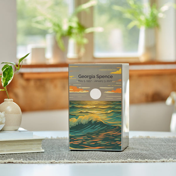 A pewter-finished stainless steel memorial urn with a vibrant ocean landscape design sits on a woven mat atop a table, with a softly lit window and green plants in the background. The artwork depicts golden waves reflecting the light of a full moon against a twilight sky. An engraving is showcased on the urn, with personalization available. This contemporary urn, available at Magnolia Cremations, offers a meaningful and artistic tribute to a loved one.