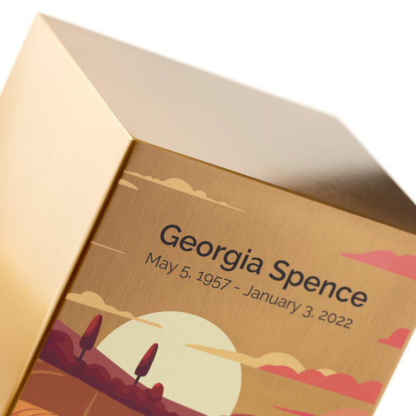 A close-up of the Prairie Landscape in Bronze Memorial Urn, available at Magnolia Cremations. The brushed bronze surface features an engraved name and dates, showcasing the option for personalization. The urn's design includes a warm, stylized illustration of a prairie at sunset, with rolling hills, hay bales, and a golden sun. The image highlights the urn's smooth, high-quality finish and detailed artwork.