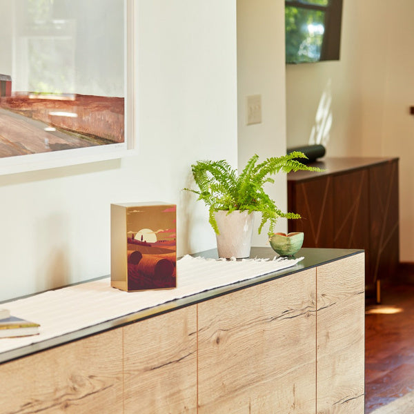A Prairie Landscape in Bronze Memorial Urn is displayed on a modern wood and black metal console table in a well-lit home setting. The urn, available at Magnolia Cremations, features a warm-toned illustration of a prairie at sunset with rolling hills, hay bales, and a golden sun. A green potted fern and ceramic bowl sit beside it, adding to the serene atmosphere. A soft, neutral-colored runner decorates the table, complementing the urn's brushed bronze finish.
