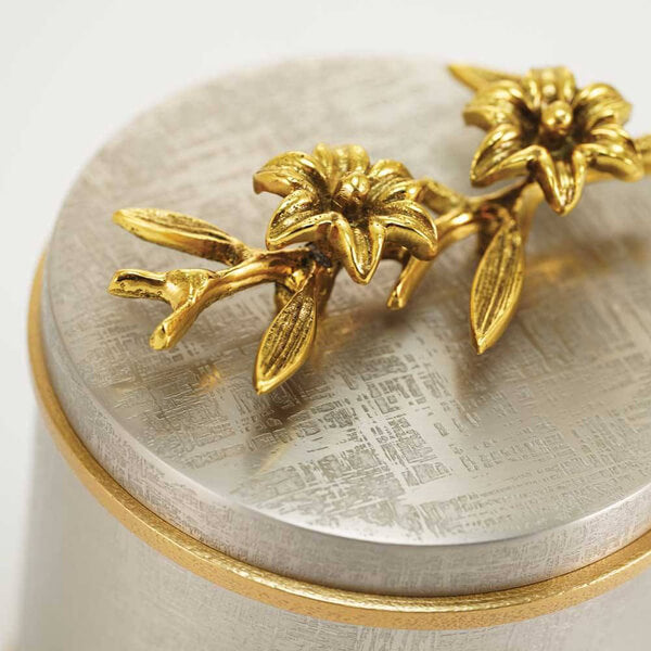 Close-up of a keepsake urn lid, showing two detailed gold sculpted flowers with leaves resting on a textured silver surface. The lid also features a gold trim around the edge.
