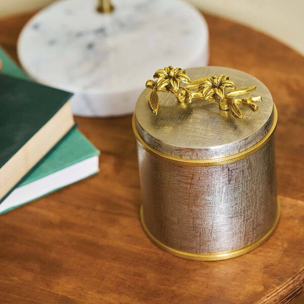 Keepsake urn with a brushed silver finish and gold trim, displayed on a wooden table. The lid is adorned with two gold sculpted flowers and leaves. Books and a white marble decor piece are nearby, creating a warm and elegant setting.