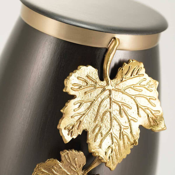 Close-up view of a slate gray cremation urn, showing detailed gold-tone vine leaves and a gold band near the lid. The surface has a smooth metallic finish and the leaf design is raised and textured.