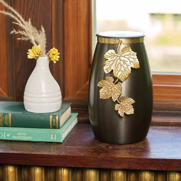 Slate gray cremation urn with a gold vine and leaf decoration displayed on a wooden windowsill. The urn is placed beside a small white vase with yellow flowers and two stacked green books, creating a warm, homey setting.