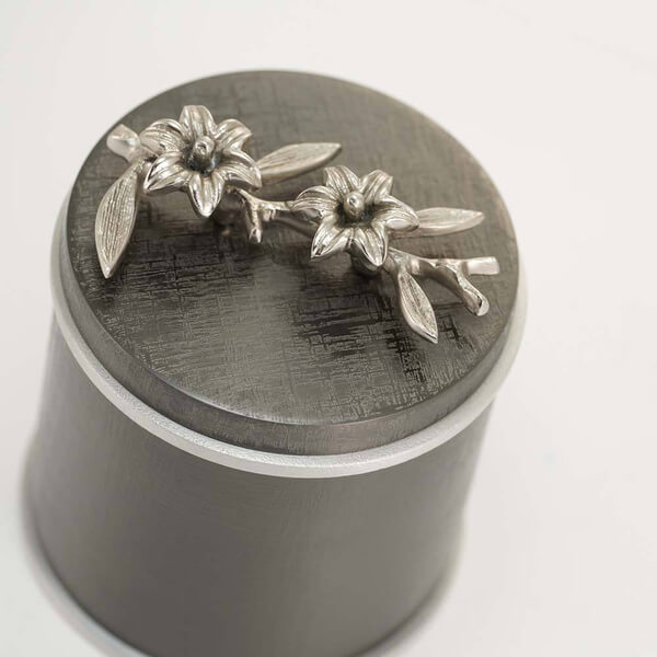 Close-up of a dark metallic keepsake urn with silver trim, showing the textured lid adorned with two detailed silver sculpted flowers and leaves.