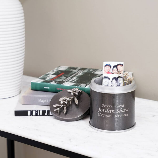 Dark metallic keepsake urn with silver floral decoration on the lid, placed on a marble surface with its lid off; optional engraving is shown on the front and personal mementos are inside.