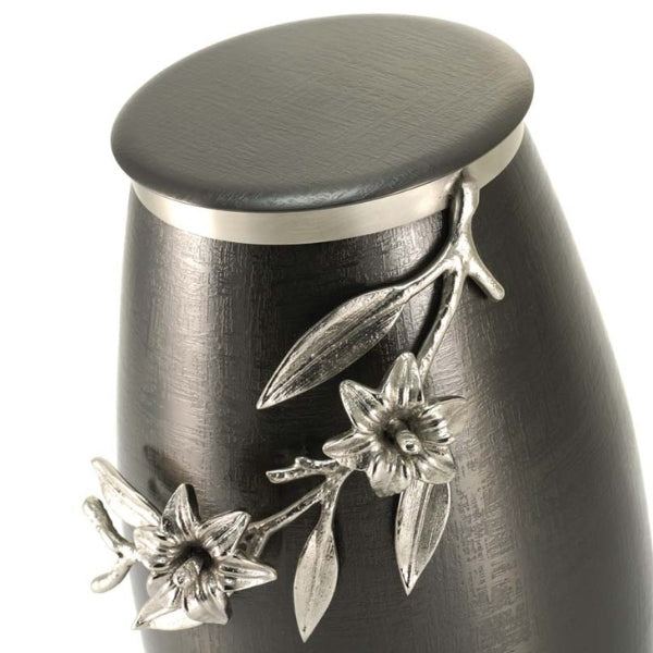 Close-up of the Windsong Lily Slate/Pewter Memorial Urn, available at Magnolia Cremations. The image highlights the intricate pewter lily branch embellishment, which is sculpted in high relief with delicate flowers, leaves, and stems wrapping around the urn’s smooth, brushed slate-black stainless steel surface. The urn’s flat stainless steel lid is visible, contrasting subtly with the textured body. The detailed floral design adds an elegant and artistic touch to the urn’s contemporary form.