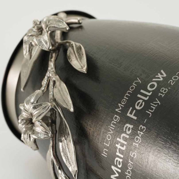 Close-up of the Windsong Lily Slate/Pewter Memorial Urn, available at Magnolia Cremations. The image highlights the urn’s textured slate-black stainless steel body with a sculpted pewter lily branch wrapping around its surface. The intricate floral design features delicate flowers, leaves, and stems in a high-relief finish. Part of an engraved inscription is visible, showcasing the personalization option available for this urn. The reflective pewter contrasts elegantly with the dark brushed metal.