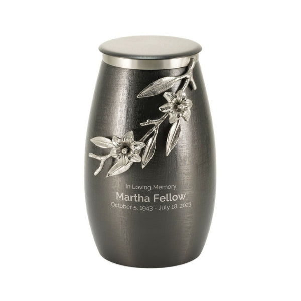 The Windsong Lily Slate/Pewter Memorial Urn, available at Magnolia Cremations. This elegantly designed urn features a brushed slate-black stainless steel body with a sculpted pewter lily branch wrapping around its surface. The delicate floral embellishment includes intricately detailed flowers, leaves, and stems in a raised metallic finish. An engraved inscription is displayed on the urn, showcasing the personalization option. The urn's contemporary shape and combination of textures create a timeless piece.